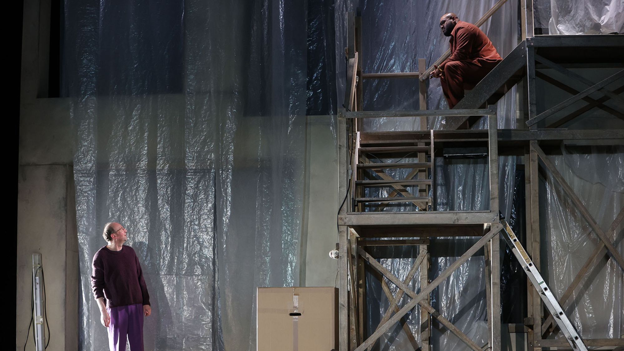 | | Ein mehrstöckiges Holzgerüst steht auf der Bühne. Ganz oben sitzt ein Schauspieler auf der Treppe. Er spricht mit einem anderen Schauspieler unten.