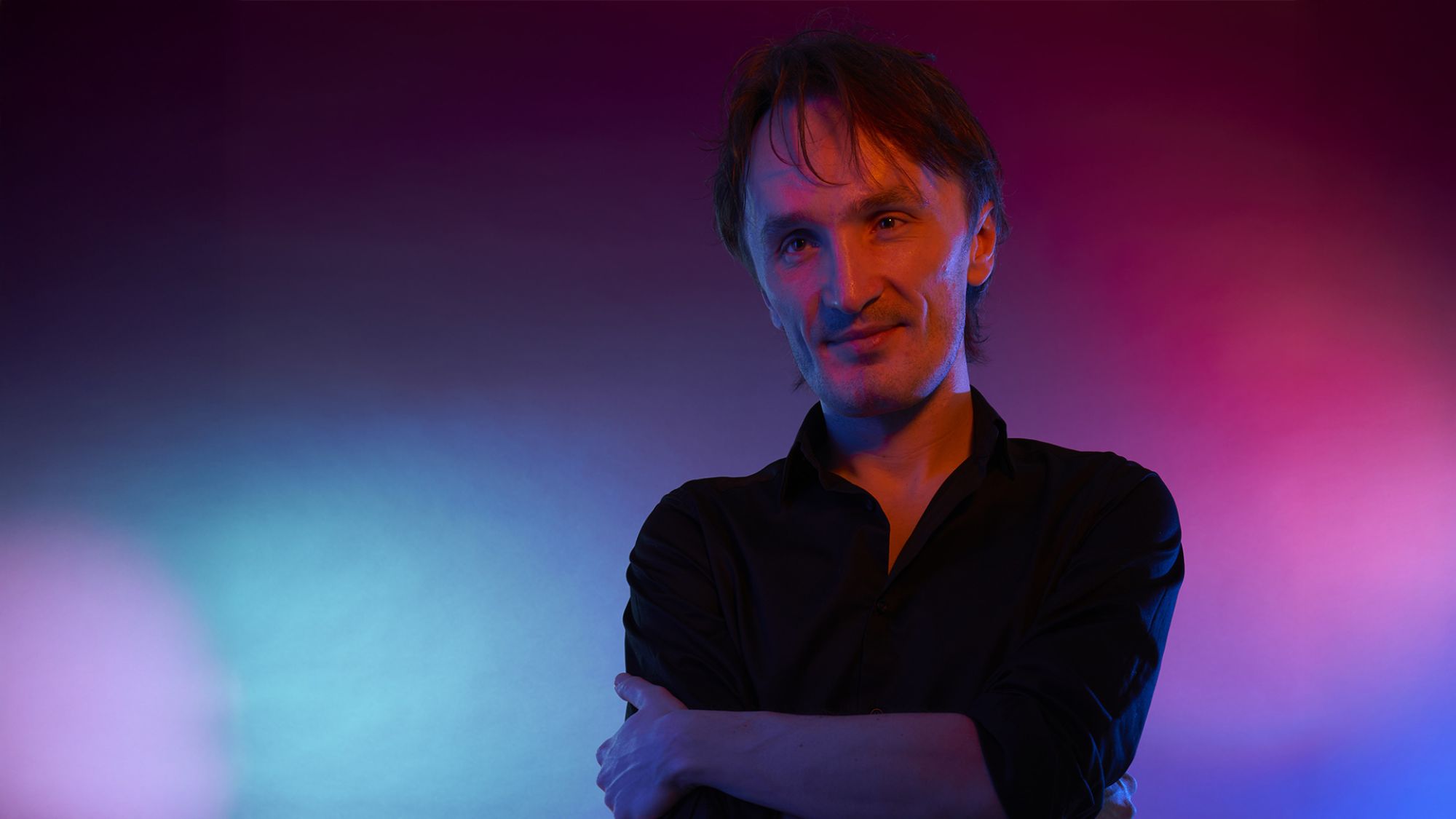 | | Ein Mann steht mit verschränkten Armen vor einem bunten Hintergrund.