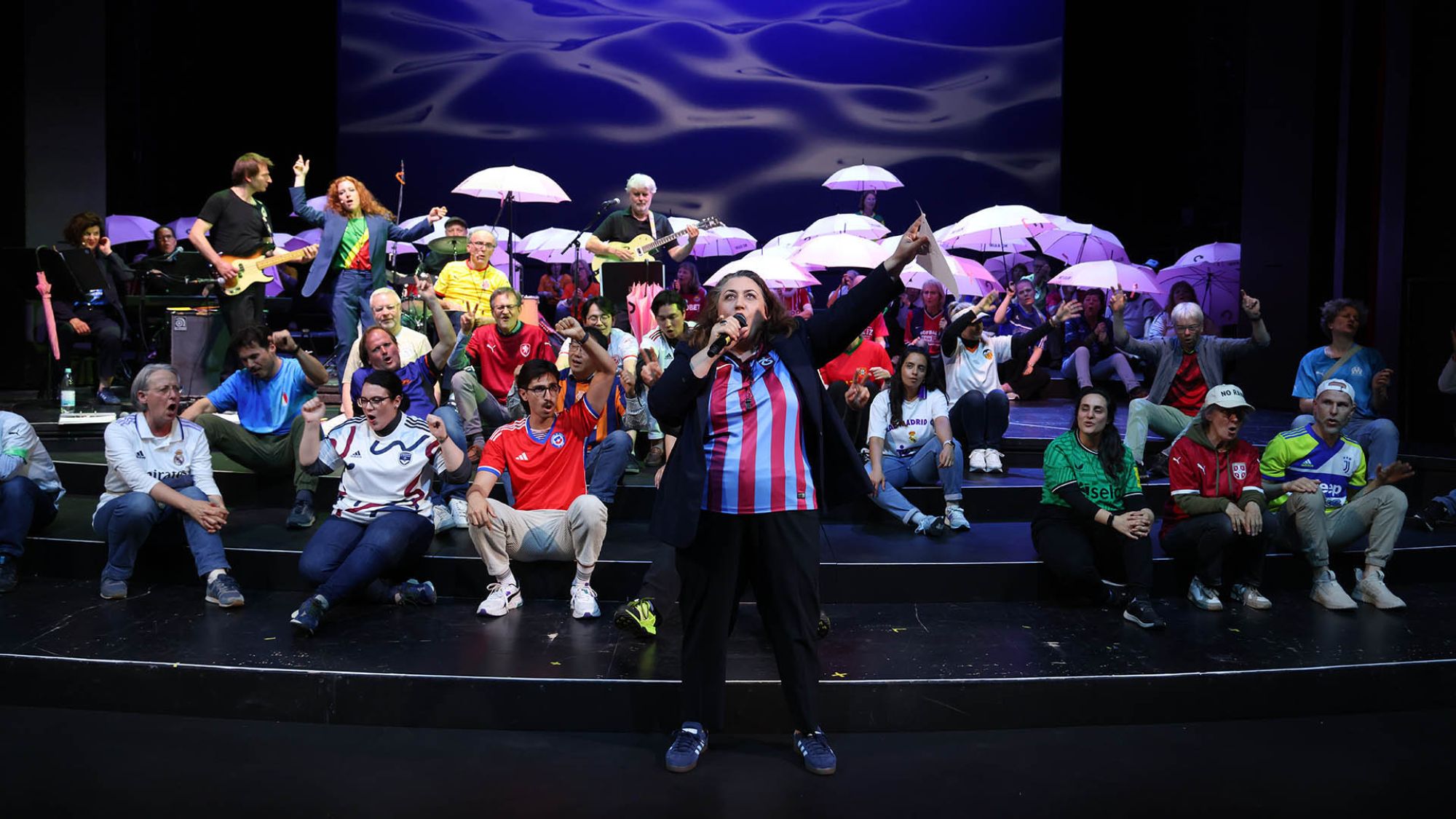 | | Auf einer Bühne sitzen viele Menschen und singen. Sie tragen Trikots in verschiedenen Farben. Vorne steht eine Frau mit Mikrofon in der Hand.