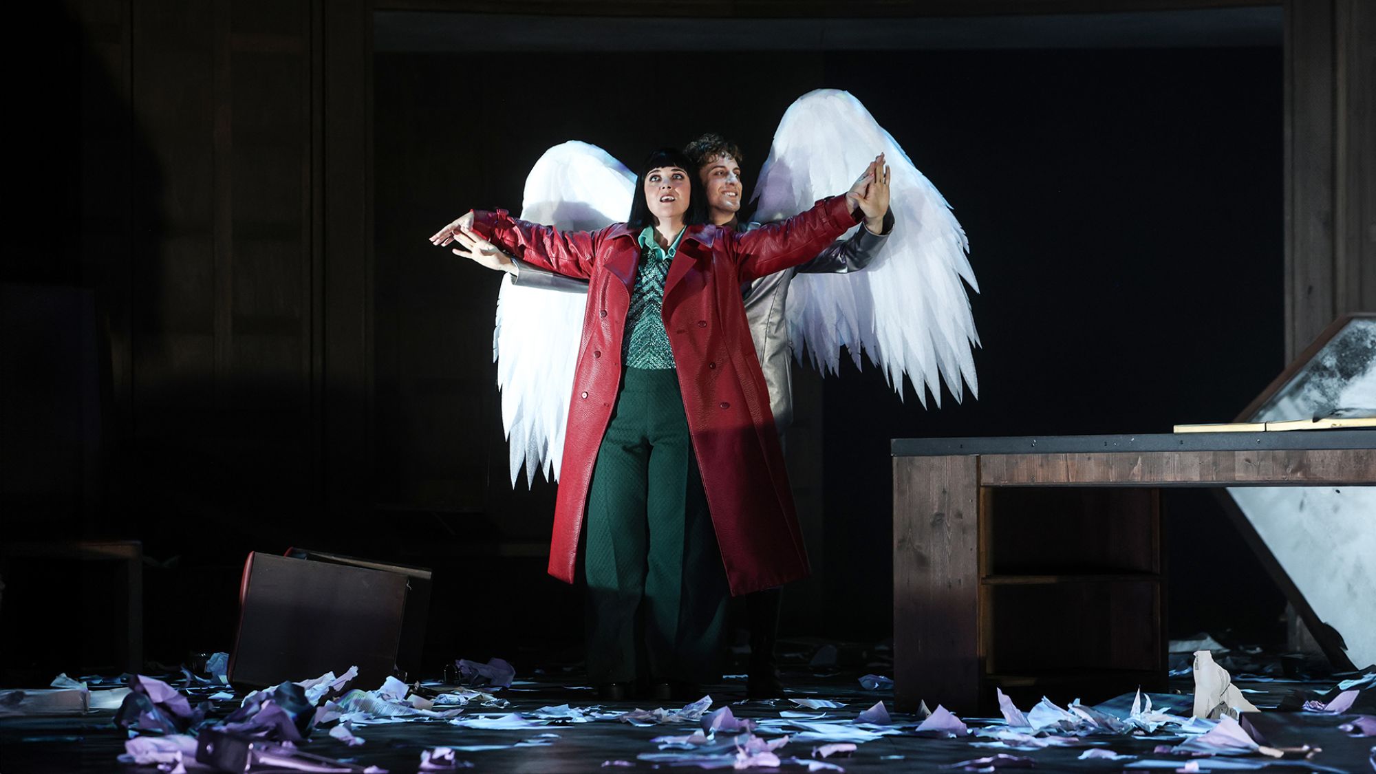 | | Eine Frau mit rotem Mantel steht auf einer Bühne. Hinter ihr steht ein Mann mit zwei großen weißen Flügeln.
