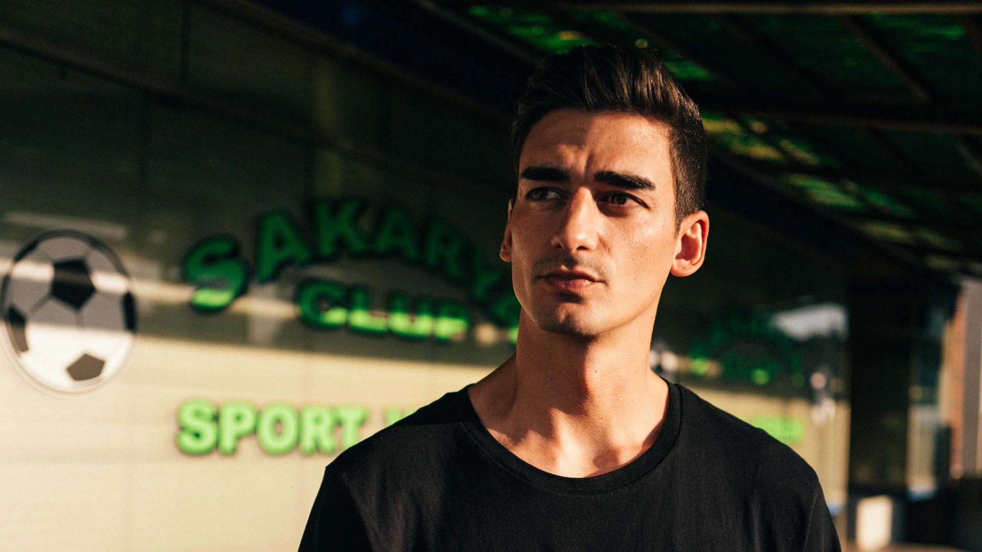 | | Robin Schimko Ein Mann mit schwarzen kurzen Haaren schaut zur Seite. Im Hintergrund ist eine Betonwand, auf der unter anderem ein Fußball aufgemalt ist.