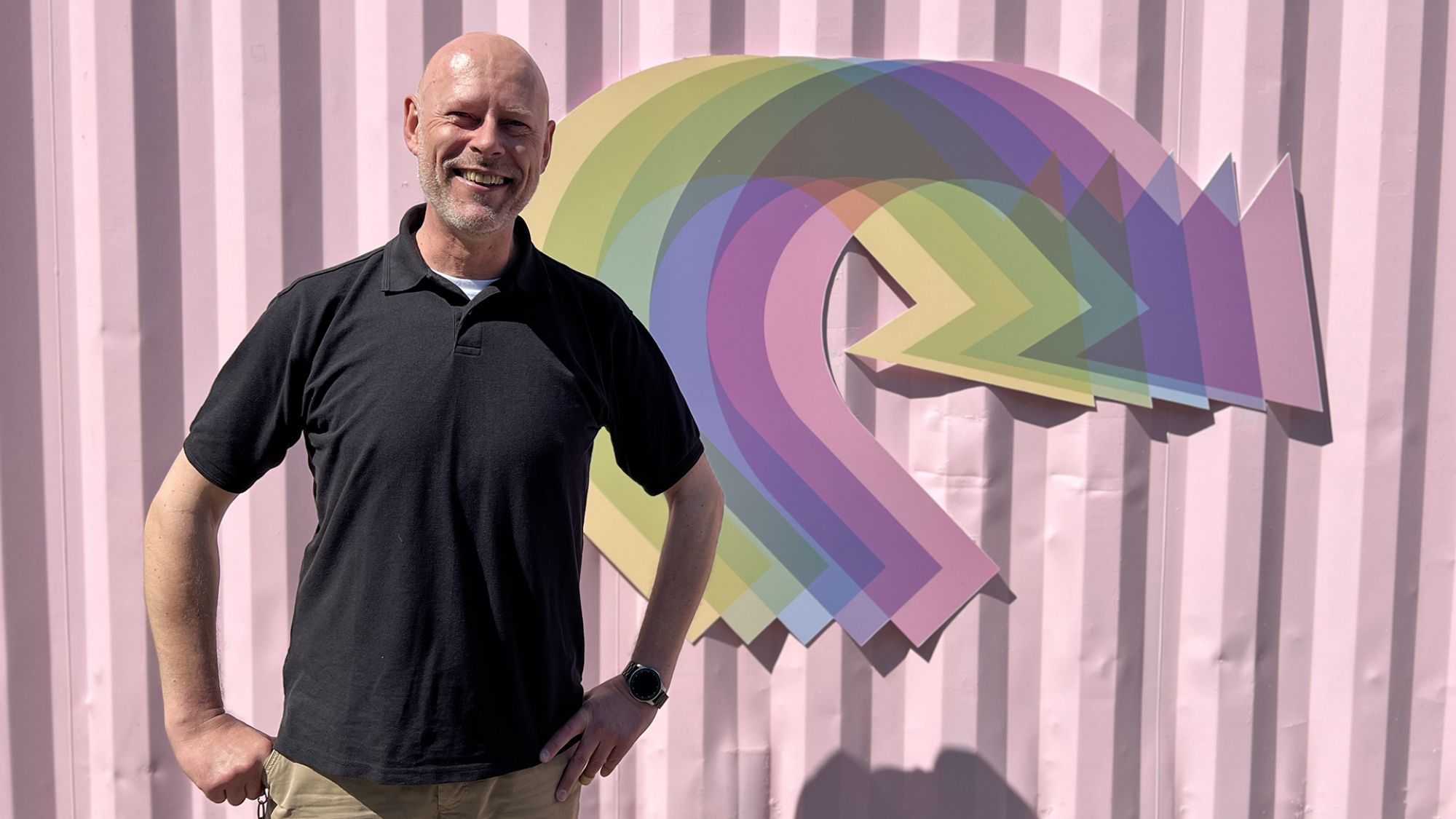 | | Ein Mann lächelt in die Kamera. Er steht vor einem pinken Container mit bunten Pfeilen.