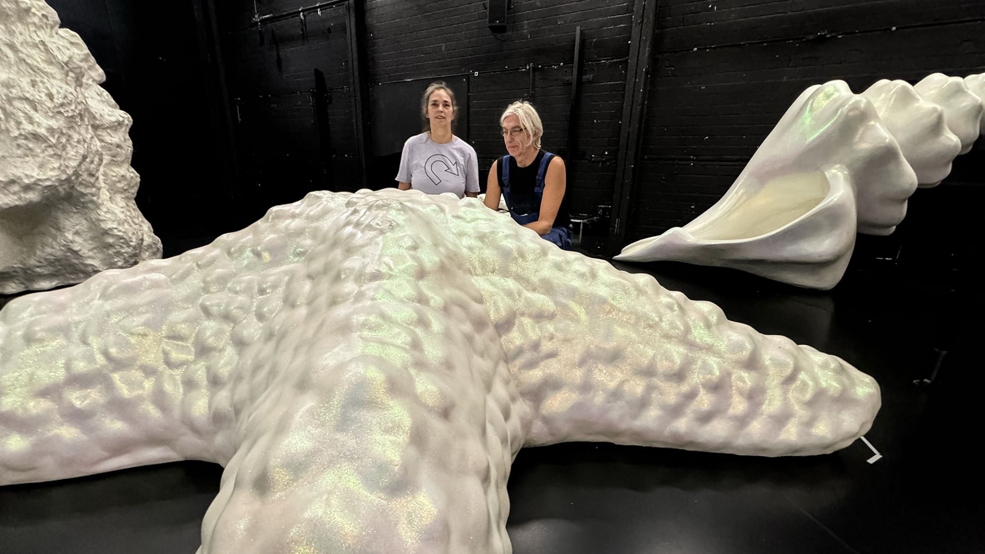 | | Auf einer Bühne liegt ein überdimensional großer weißer Seestern. Hinter dem Seestern knien zwei Menschen.