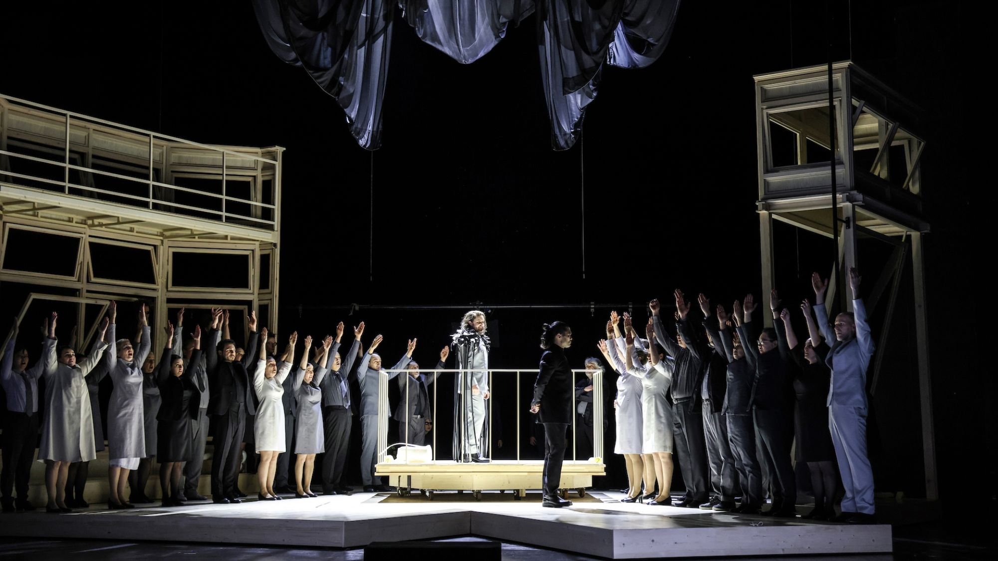 | | In der Mitte der Bühne ist eine umgitterte Erhebung. Auf ihr steht ein Sänger. Der ganze Opernchor steht zu beiden Seiten. Alle halten die Arme hoch.