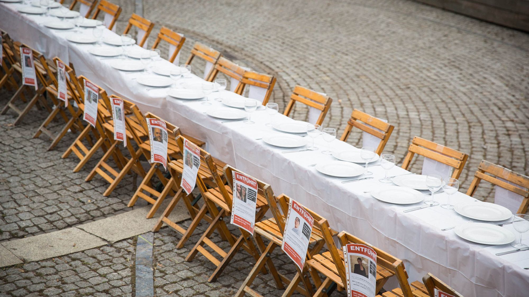 | | Jasper Wessel Auf einem Platz steht ein langer Tisch mit weißer Tischdecke und leeren Tellern. An den Stühlen hängen Zettel. Auf den Zetteln steht in Großbuchstaben entführt. Darunter sind Fotos von verschiedenen Personen abgebildet.