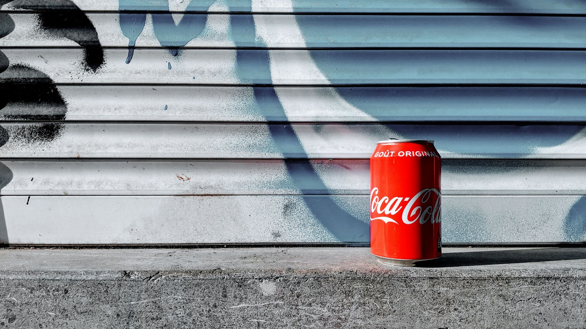 | | Lucian Alexe / Unsplash Eine Cola-Dose steht vor einem blau-weißen Rolltor.