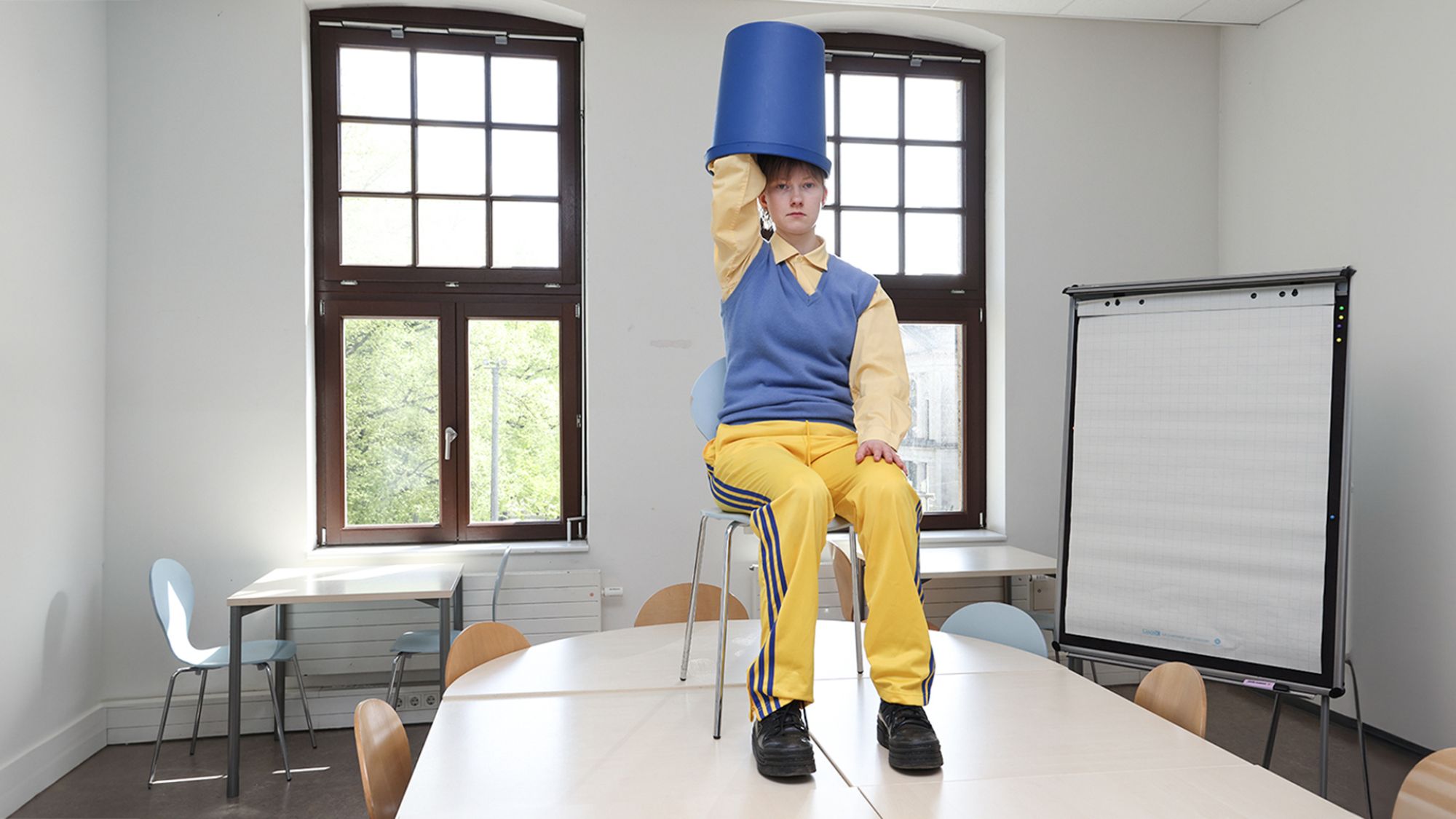 | | Eine Person sitzt mit einem Stuhl auf einem Tisch. Auf ihrem Kopf trägt sie einen umgedrehten Mülleimer.