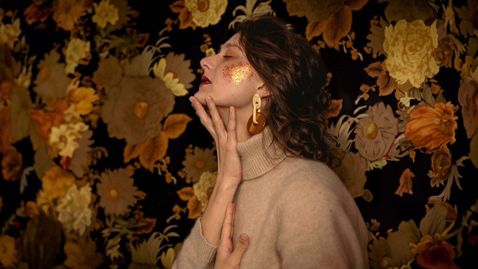| | Marie Fady Eine Frau steht vor einer Tapete mit Blumen. Sie hat die Augen geschlossen und hält ihre Hand ans Kinn. Auf ihrer Wange ist goldener Glitzer.