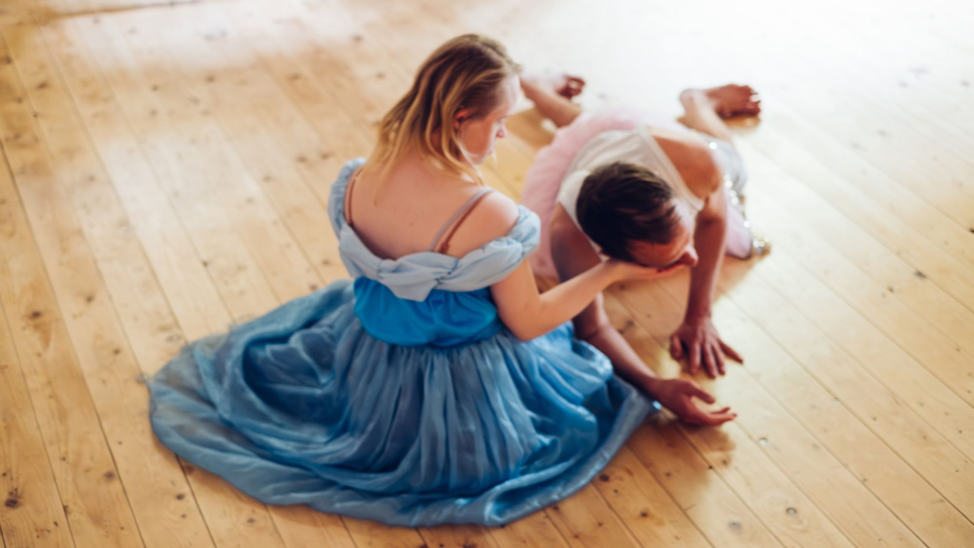 | | Daniela Buchholz Eine Frau mit einem hellblauen Kleid sitzt auf einem Holzboden. Neben ihr liegt ein Mann. Er legt sein Gesicht in ihre Hand.