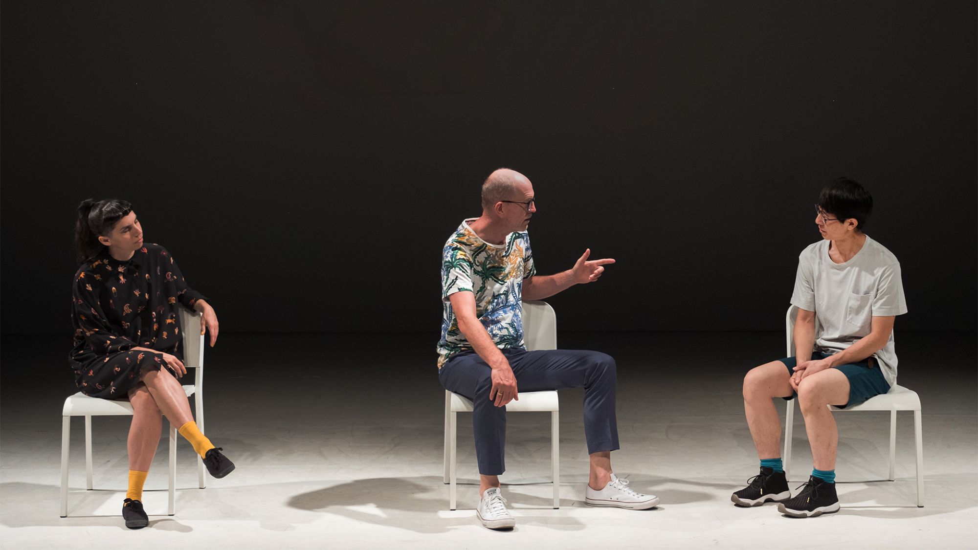 Drei Personen sitzen auf Hockern auf einer Bühne.