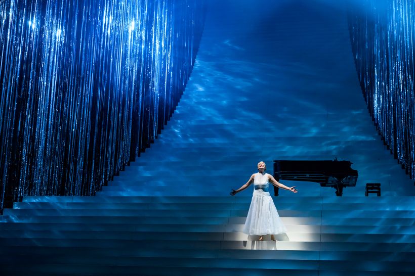 © Bettina Stöß Eine Frau in einem weißen Kleid steht auf einer beleuchteten Bühne mit blauem Licht und einem schwarzen Klavier im Hintergrund.