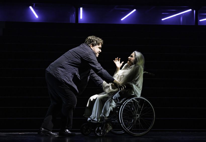 © Bettina Stöß Eine Person im Rollstuhl wird auf einer dunklen Bühne von einer anderen Person dramatisch angesprochen, Neonlicht im Hintergrund.