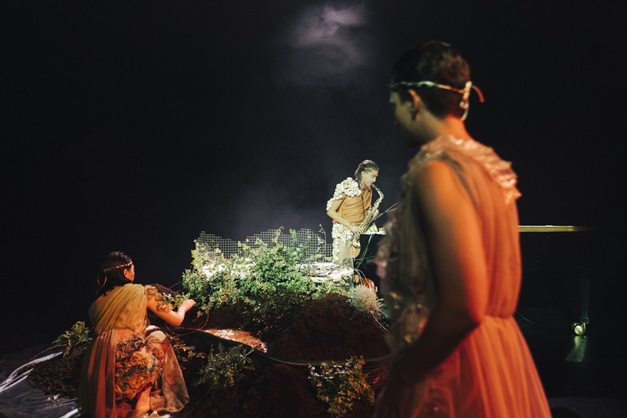 Der Musiker Ralph Heidel steht Saxofon spielend auf einem Hügel aus Farnen, Gräsern und Moosen. Ein junger Mann in Toga blickt auf eine junge Frau, die unterhalb des Hügels den Musiker beobachtet.