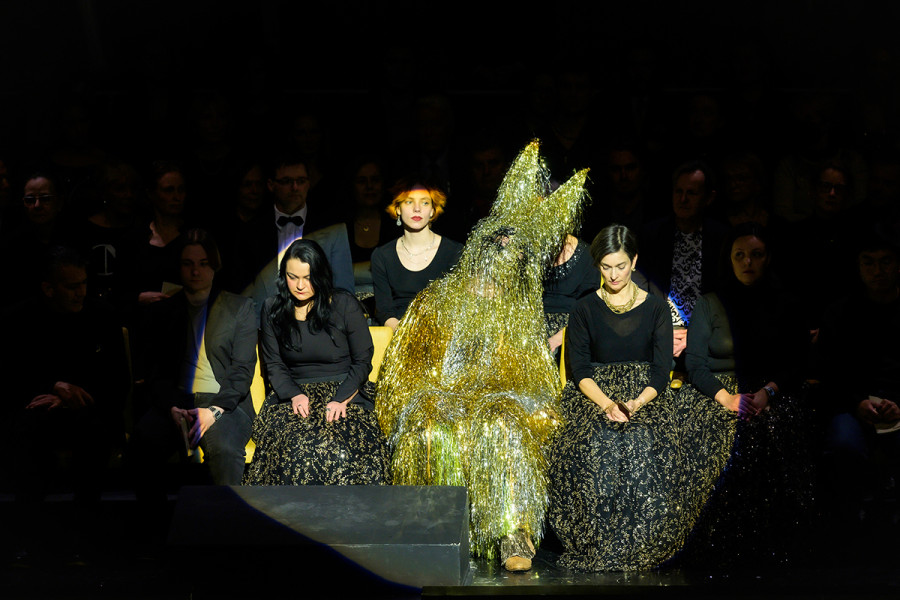 Rigoletto sitzt im Goldlametta-Hasenkostüm in den Stuhlreihen des Eröffnungsbühnenbildes. Der Saal der Deutschen Oper Berlin mit seinen typischen gelben Sitzbezügen ist mit Parkett und erstem Rang gespiegelt. In diesem zweiten Saal sitzen Chor und Statisterie.