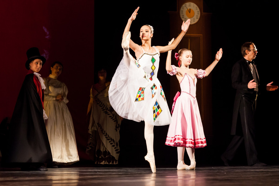 Im Vordergrund tanzt eine Elevin der Kinder Ballett Kompanie in einem Columbine-artigen Kleid mit bunten Rauten auf Mieder und Tütü.