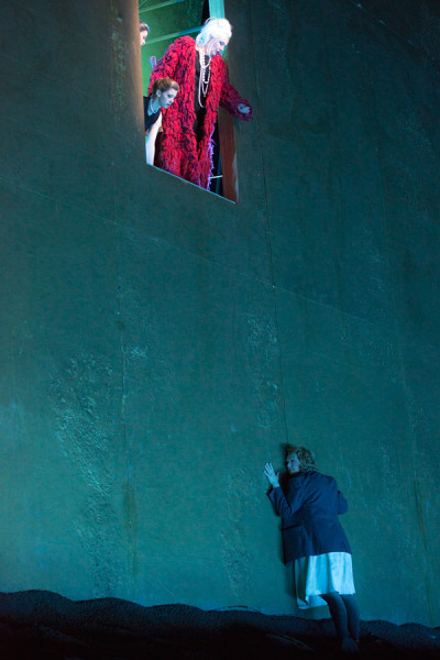 Elektra lehnt an der Rückwand, die in grelles Grün getaucht ist. Im oberen Teil der Wand hat sich eine Tür geöffnet, aus der heraus Klytämnestra auf sie herabblickt.