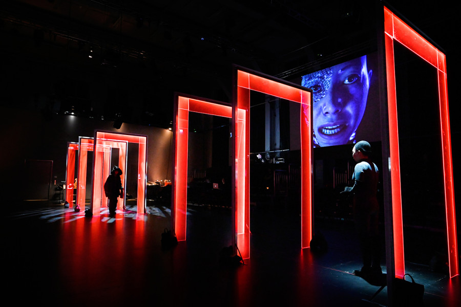 Dieses Bild zeigt eine Bühnentotale. Perspektivisch nach hinten stehen die rotbeleuchteten Türen. Im Hintergrund der Orchesteraufbau in warmem orangenen Licht, im vorderen Teil des Bildes das Avatar Lou.