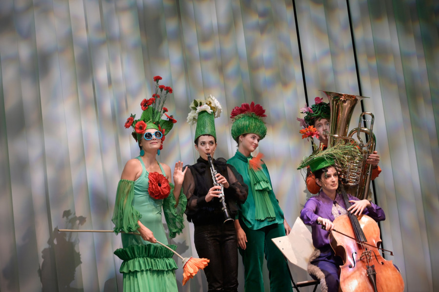 Die Blumenkönigin trägt ein grünes Trägerkleid mit Volants. Alle Akteure tragen Blumenhüte. Neben der Königin die Instrumentalsolist*innen mit Klarinette, Cello und Tuba.