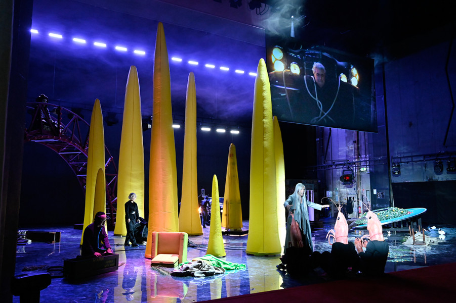 Pilze und riesiger Garnelen bevölkern die Bühne. Auf dem Bildschirm Henry Kissinger, unten in einem silbernen Anzug Chou En-lai.