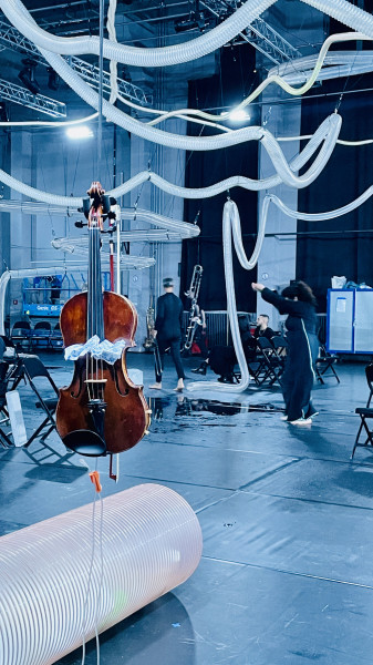 Eine Impression aus SHALL I BUILD A DAM: Ein Cello hängt im Bühnenboden. Auf dem Boden, in der Technik Schläuche.