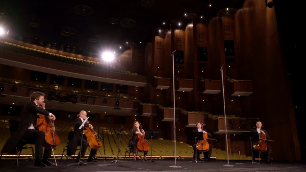 Dieses Bild ist das Standbild zu einem kammermusikalischen Video, in dem fünf Cellisten Highlights aus DON CARLO interpretieren. Die fünf Musiker*innen sitzen mit dem Rücken zum Saal mit leeren Stuhlreihen.
