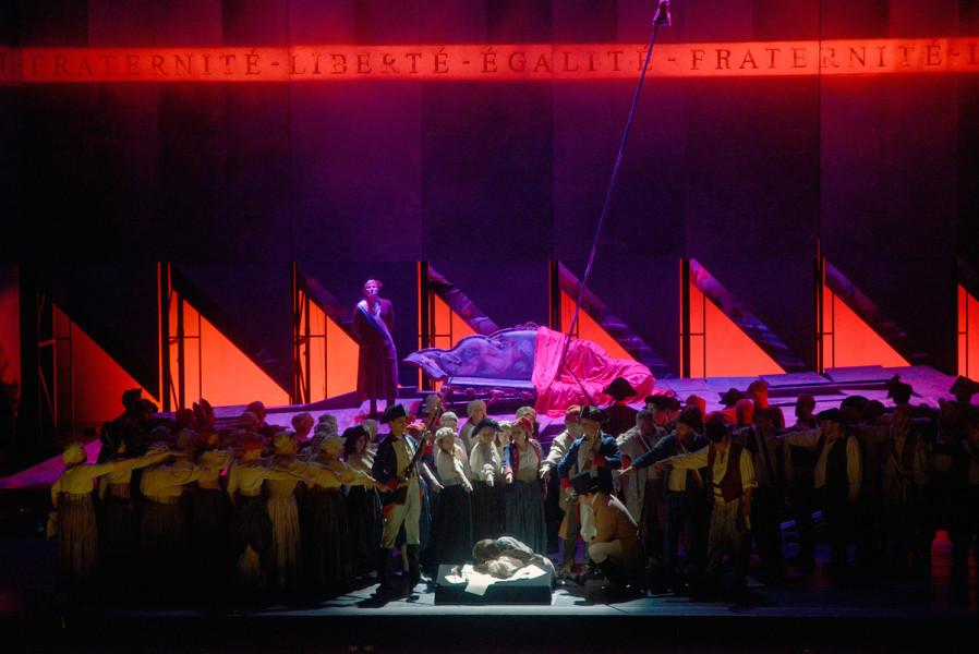 Paris brennt. Grell angeleuchtete Bühnenbildteile leuchten in tief rot. Das Volk von Paris verurteilt Andrea Chenier. Rings um die Bühne läuft ein großes Spruchband: Liberté, Égalité, Fraternité sind "in Stein gemeißelt". 