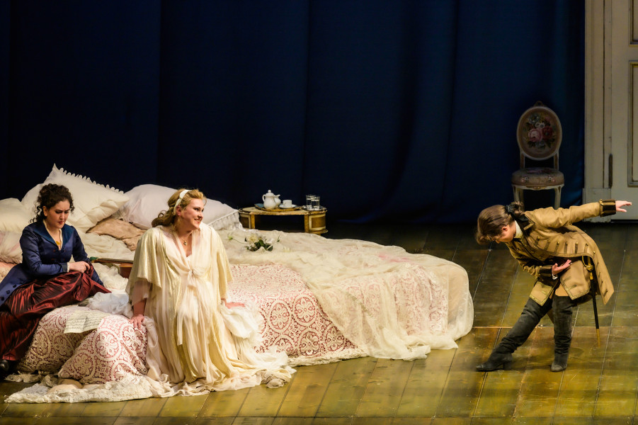 Susanna und die Gräfin sitzen in der Gräfin Schlafgemach auf dem Bett. Cherubino kommt mit einer tiefen Verbeugung ins Schlafgemach.