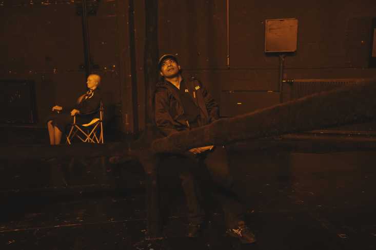 Ein schwarz gekleideter Mann lehnt an einer Wand. Hinter ihm sitzt eine lebensgroße Puppe auf einem Klappstuhl.
