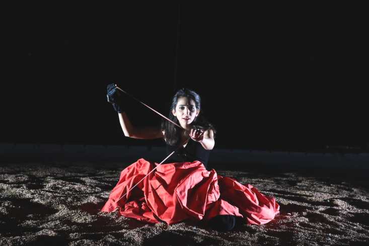Die Sängerin sitzt in einem großen, roten Rock auf dem Boden im Sand. 