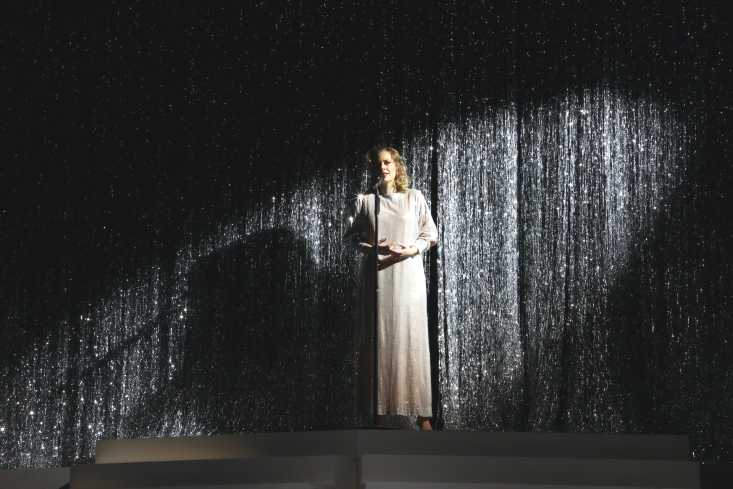 Eine Sängerin steht vor einem Lamettavorhang. Sie trägt ein langes weißes Kleid und steht im Scheinwerferlicht.