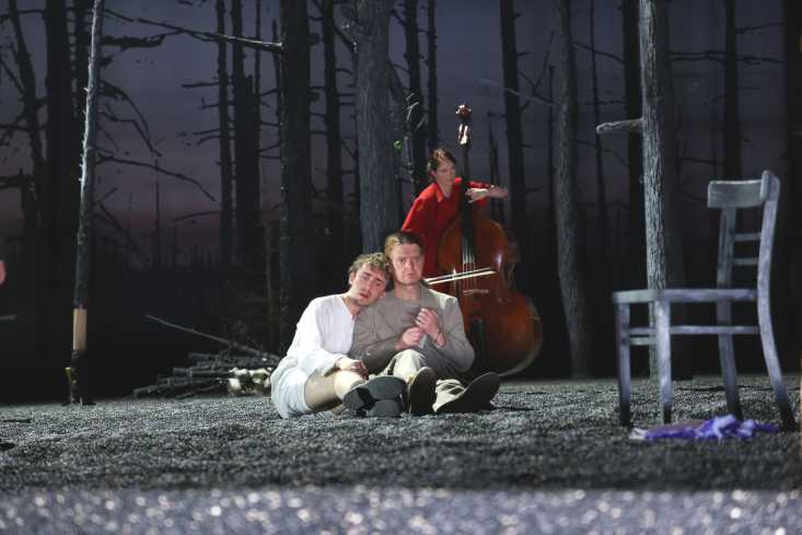 Ein Mann sitzt an einen Baum gelehnt auf dem Boden. An ihm lehnt ein anderer Mann. Hinter den beiden steht eine Frau in einem roten Kleid und spielt Kontrabass.