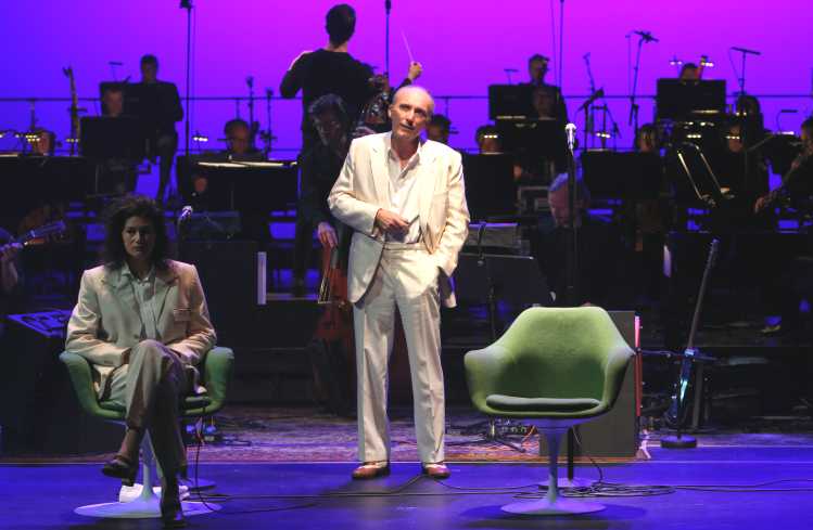 Auf der linken Bildseite sitzt eine Frau in einem weißen Anzug auf einem Drehsessel. Neben ihr steht, ebenfalls ganz in weiß, ein Mann. Im Hintergrund spielt das Orchester, der Dirigent steht am Pult.