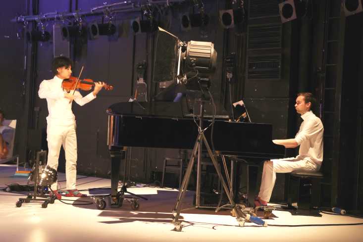 Zwei Musiker. Der eine sitzt an einem Flügel, der andere spielt Geige. Beide sind komplett weiß gekleidet.