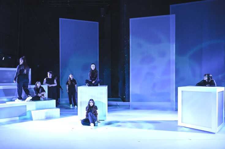 © Manja Herrmann // Eine Großaufnahme der Bühne. Links ist eine große Treppe, in der Mitte und rechts steht jeweils ein Kubus. Die Bühne ist blau beleuchtet. Die Spielerinnen stehen und sitzen darin verteilt.