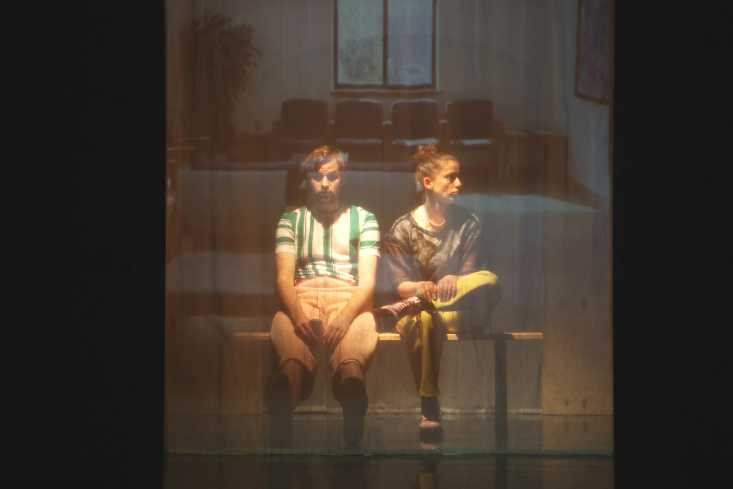 Zwei Personen sitzen auf einer Bank nebeneinander. Im Hintergrund ist ein Wartezimmer. 