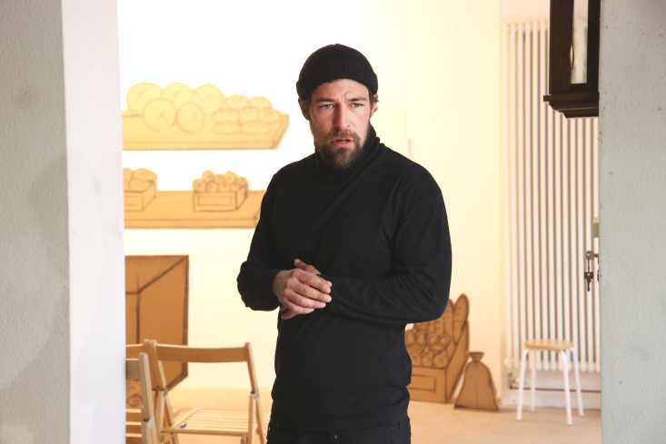 Ein Mann steht schwarz gekleidet in einem Raum. An der Wand hinter ihm hängen Pappbilder. Auf ihnen sind verschiedene Backwaren wie Brötchen und Kuchen gemalt. 