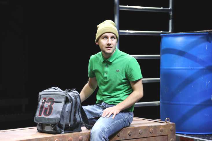 Ein Schauspieler sitzt mit Mütze und Schultasche auf einer Kiste.