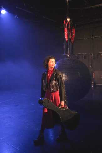 Eine Frau mit rotem Kleid und Westernjacke geht mit einem Gitarrenkoffer in der Hand über die Bühne. Hinter ihr steht eine andere Frau auf einer großen über der Erde schwebenden Kugel.