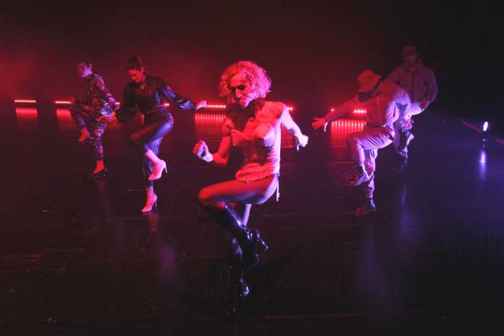 Fünf Menschen tanzen synchron in einer Formation auf der Bühne. Die Spitze bildet ein Mann mit einer blonden Perücke in einem Madonna-Outfit. Er trägt hochhackige Stiefel mit Netzstrümpfen, dazu ein Mieder mit Strumpfbändern, die lose herumfliegen. 