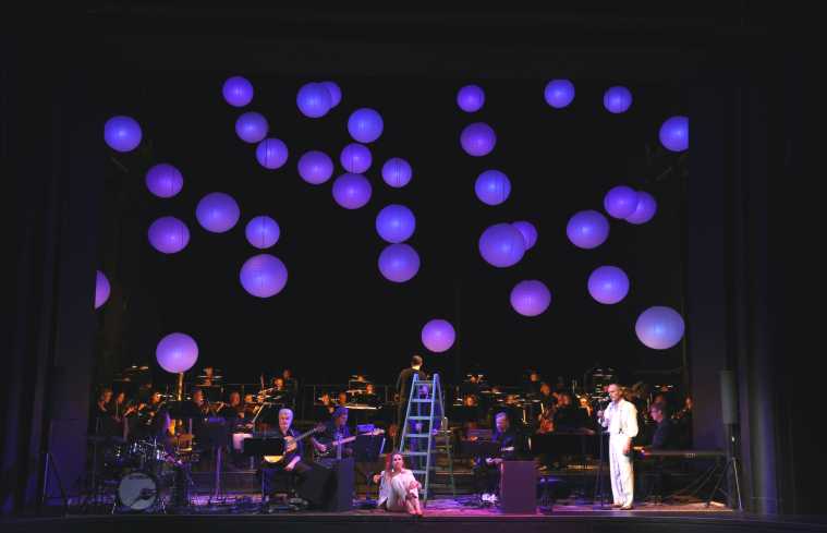 In der Mitte der Bühne sitzt eine Sängerin auf dem Boden, rechts neben ihr steht ein Mann und singt. Im Hintergrund spielt eine Band und das große Orchester. Vom Bühnenhimmel hängen sehr viele violett leuchtende Lampions.