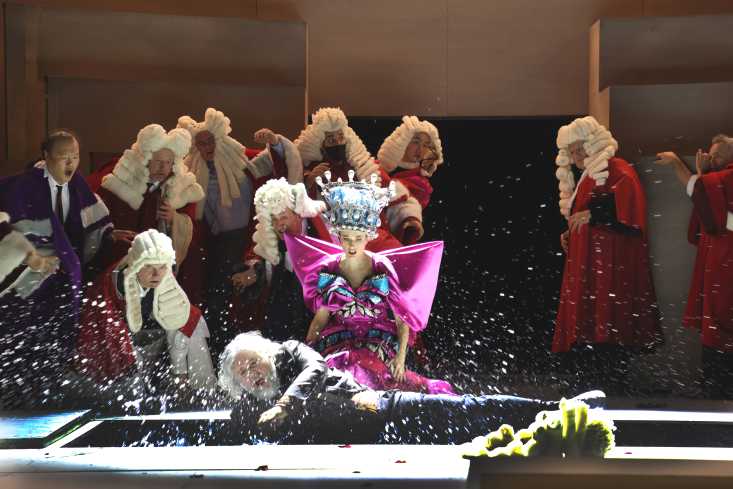 Ein Sänger landet vorn im Wassergraben. Hinter ihm kniet eine Frau in einem grellbunten Königinnenkostüm, dazu eine Gruppe von Richtern mit Roben und Perücken.