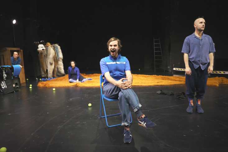 Ein Schauspieler sitzt auf einem Klappstuhl in der Bühnenmitte. Sein Mund ist aufgerissen. Die drei anderen sind hinter ihm verteilt.