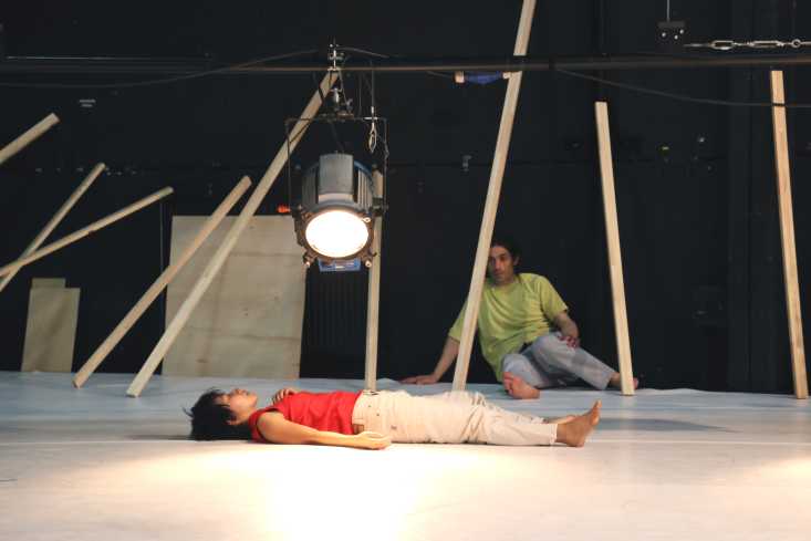 Eine Tänzerin liegt auf dem Rücken auf dem Boden. Nah über ihr leuchtet ein Scheinwerfer. An der Wand hinten sitzt ein Tänzer.