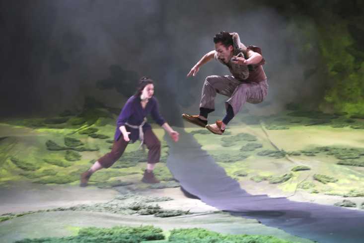 Ein Schauspieler springt und ist mitten im Flug hoch in der Luft. Eine Schauspielerin nimmt Anlauf um ebenfalls zu springen.