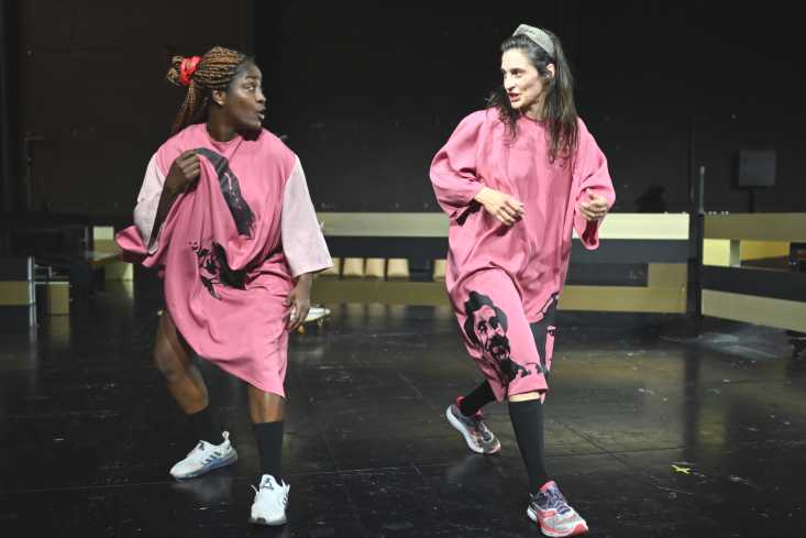 Zwei Performerinnen in rosanen Gewändern tanzen nebeneinander. Sie schauen sich an. 