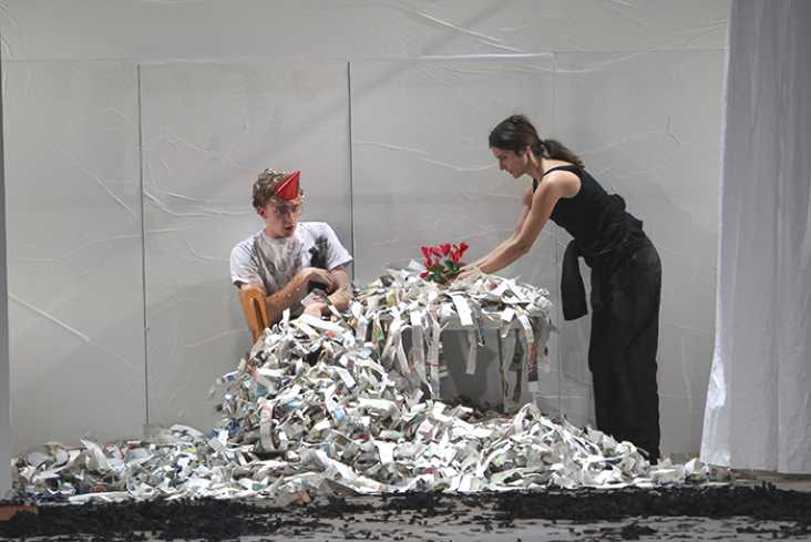 Ein Mann sitzt mit einem Partyhütchen auf dem Kopf an einem Tisch. Eine Frau stellt Blumen auf den Tisch. Alles ist bedeckt mit großen Zeitungsfetzen.