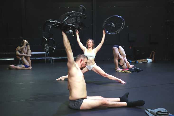 Acht Tänzerinnen und Tänzer sind auf der Bühne verteilt. Ein Tänzer sitzt vorn auf dem Boden und hält einen Teil eines Rollstuhls in die Höhe, hinter ihm hält eine Tänzerin die Räder des Rollstuhls.