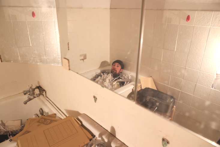Ein Mann liegt angezogen in einer Badewanne voller Papierschnipsel. Er blickt in den Spiegel. 