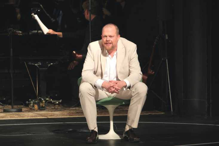 Ein Mann in einem weißen Anzug sitzt mit den Ellbogen auf den Knien nach vorn gebeugt in einem Drehstuhl und schaut ins Publikum.