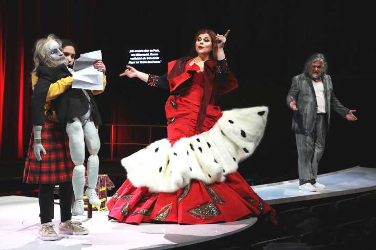 Ein Mädchen steht mit einer Falstaff-Puppe auf einem Steg im Zuschauerraum. Vor ihm eine Frau in einem üppigen roten Kleid. Hinter ihr steht der Sänger, der Falstaff spielt.